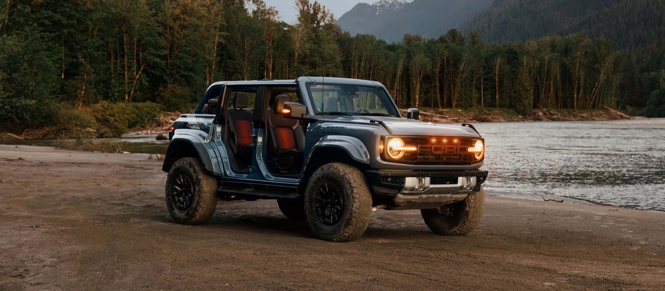 A 2025 Ford Bronco® SUV with the doors off and lights on, parked near a lake in the forest
