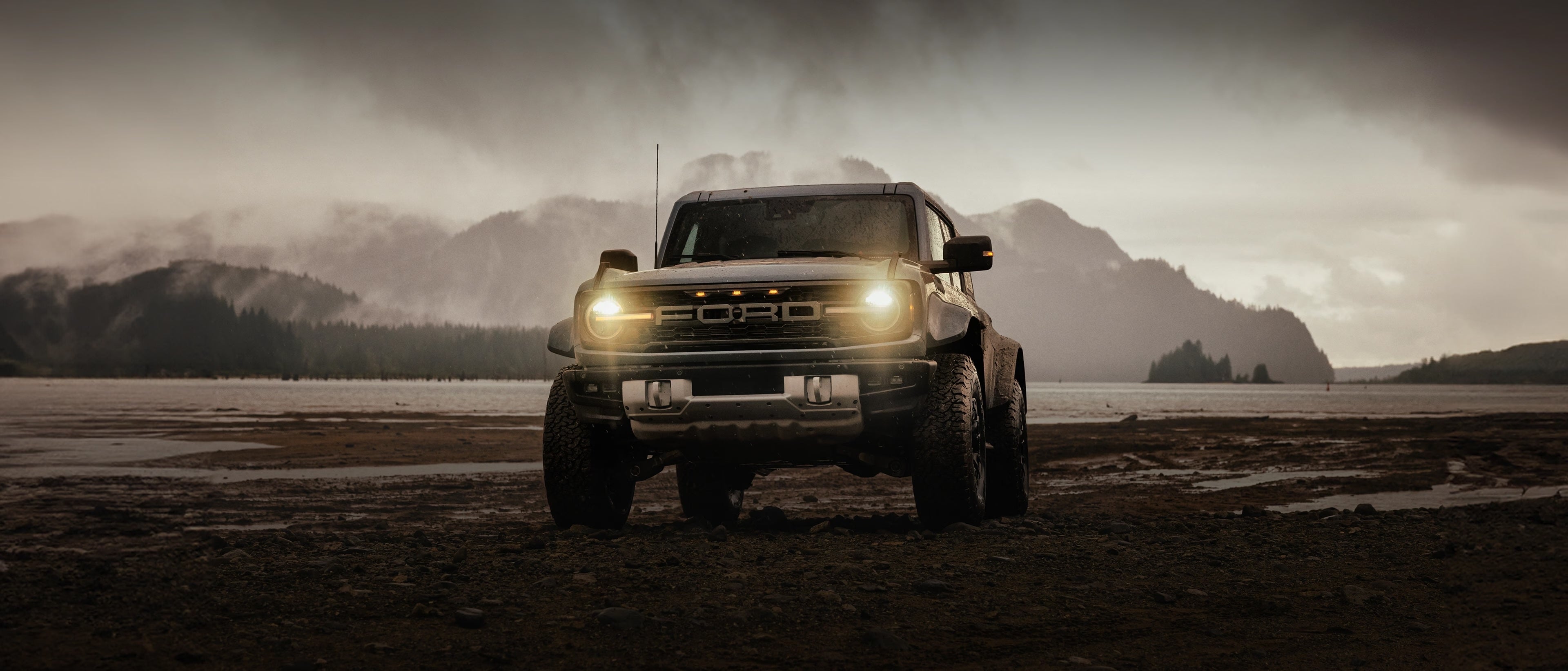 A 2025 Ford Bronco® SUV on a mud flat near a snowcapped mountain, with rain clouds threatening